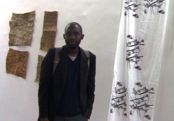 Meni Mbugha entre les réalisations pygmées en écorces battues et un pan de tissu aux motifs inspirés de l’art pygmée